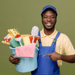 smiling holding and points at bucket of cleaning tools young africanamerican cleaner male in uniform with gloves isolated on green background